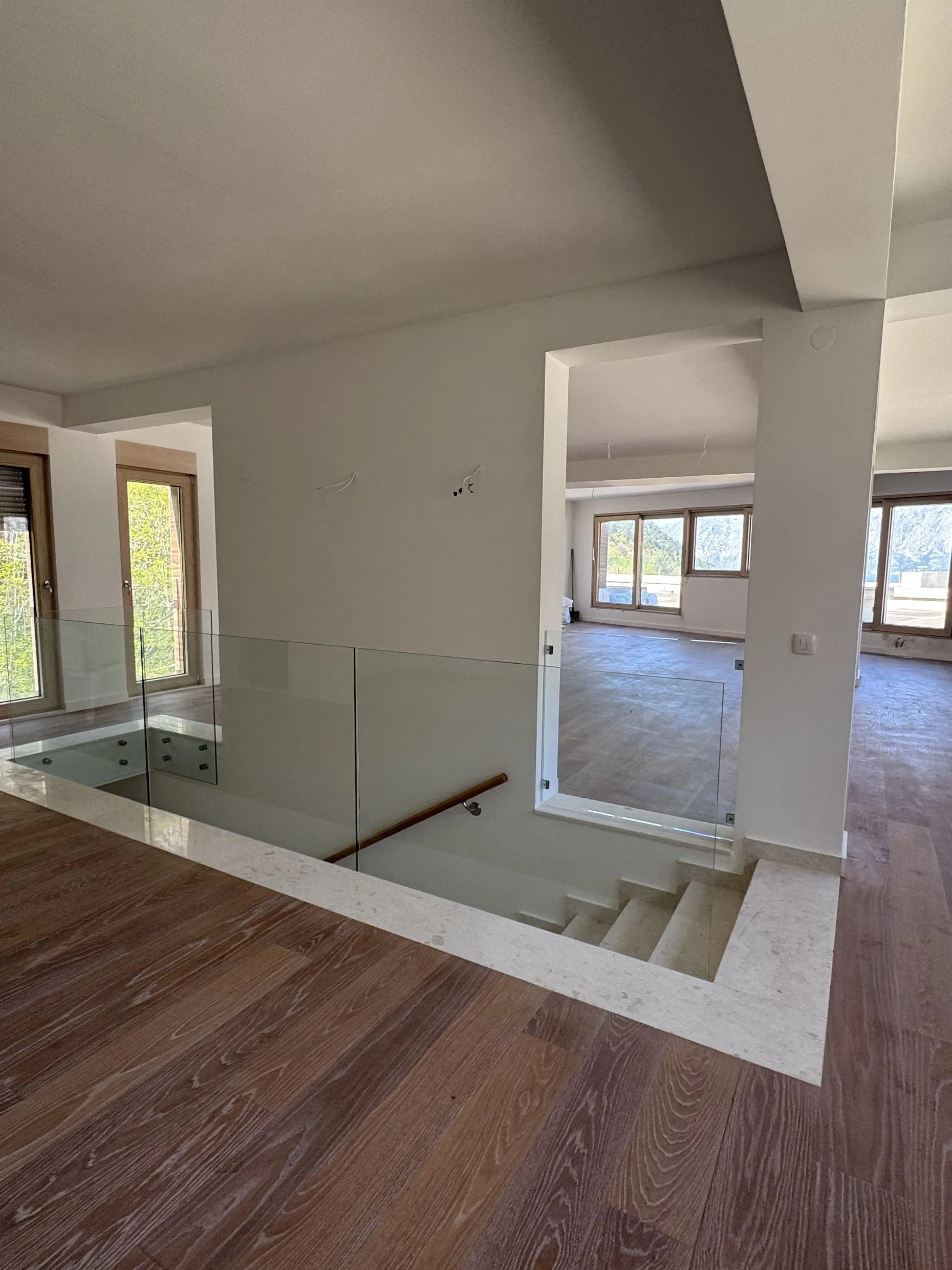 Second floor hallway with wooden flooring and glass staircase railing. White walls, doorways, and windows are visible in the background.