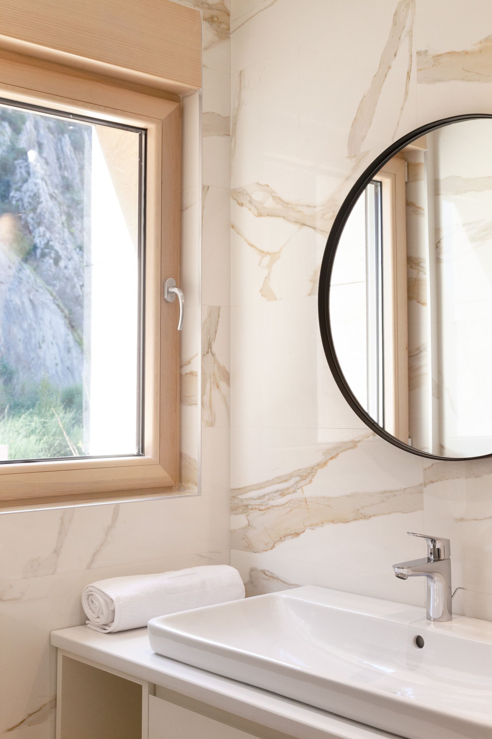 Modernes Badezimmer in einer Wohnung in Kotor, Montenegro: helle Fliesen in Marmoroptik, runder Spiegel und Blick auf die Berge.