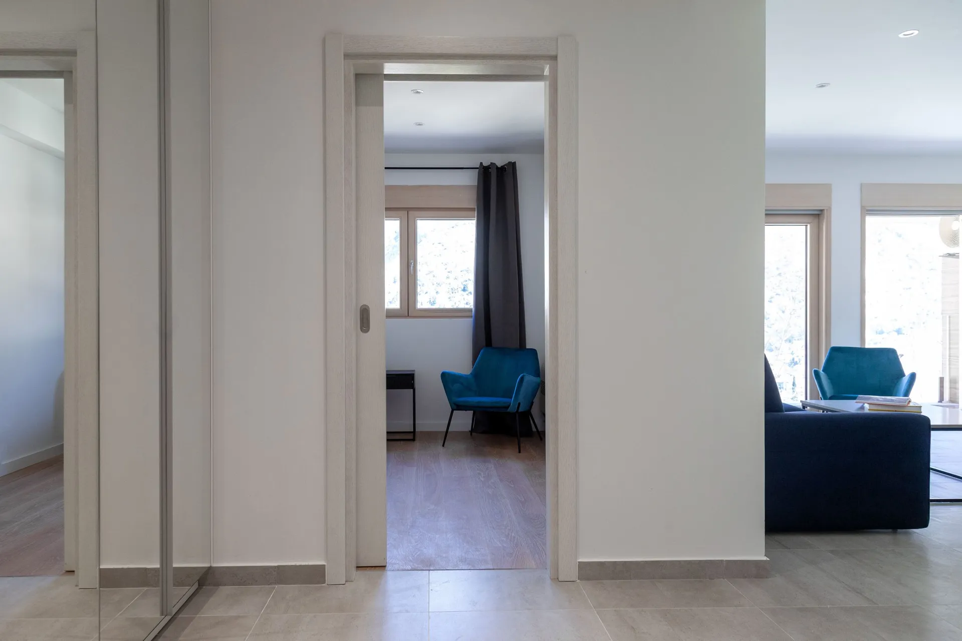 Modern living room with a blue armchair by the window and light walls, viewed from the hallway.