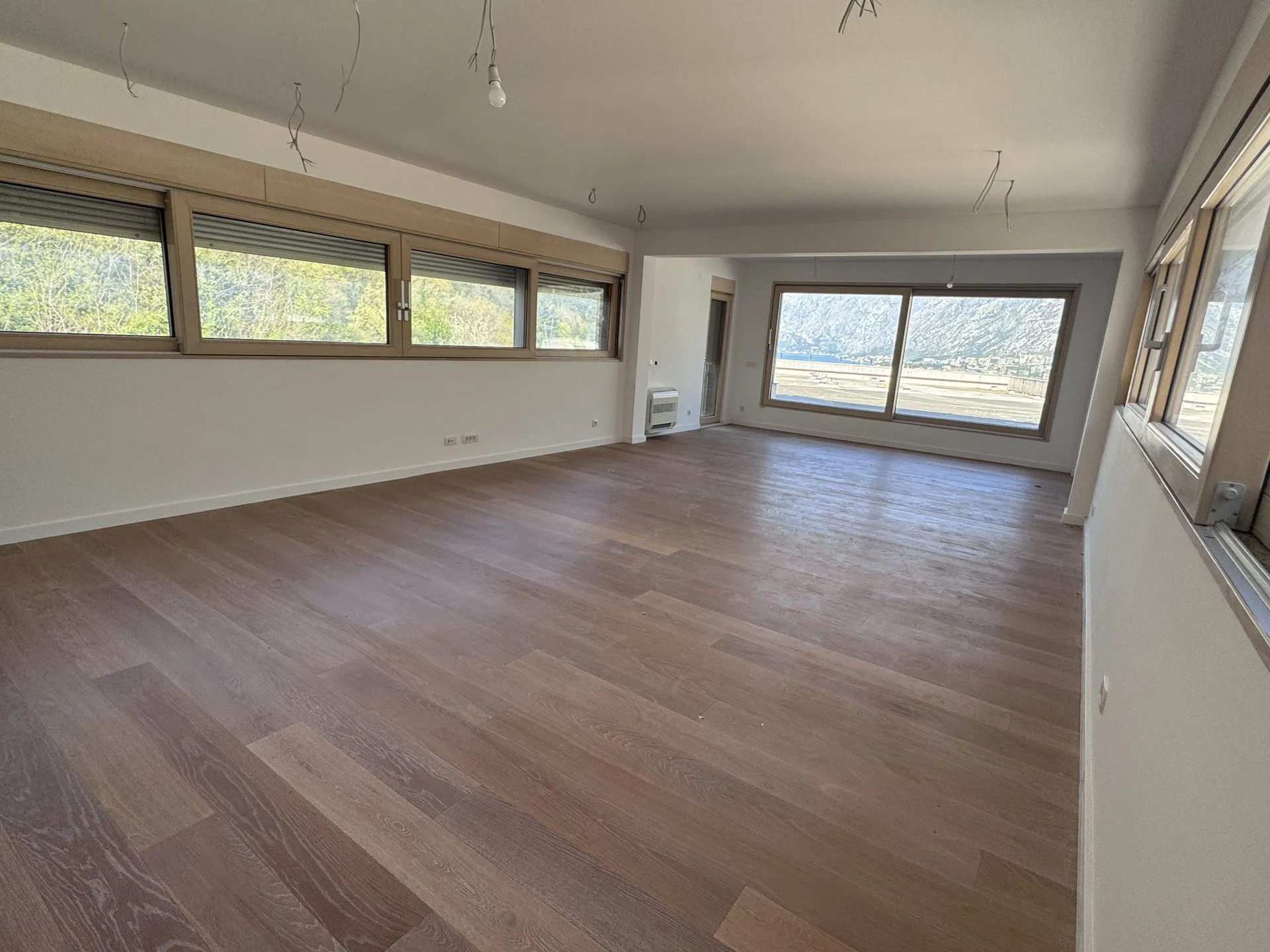 Leerer Raum mit Holzboden und großen Fenstern. Sichtbare Stromkabel für Beleuchtung und Bergpanorama.