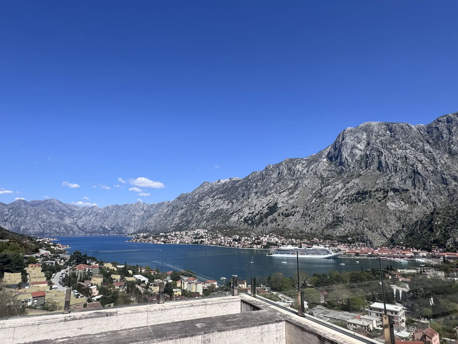 Blick von einer Terrasse auf eine Bucht, Berge und Stadt. Ein großes Kreuzfahrtschiff ist im Wasser sichtbar.
