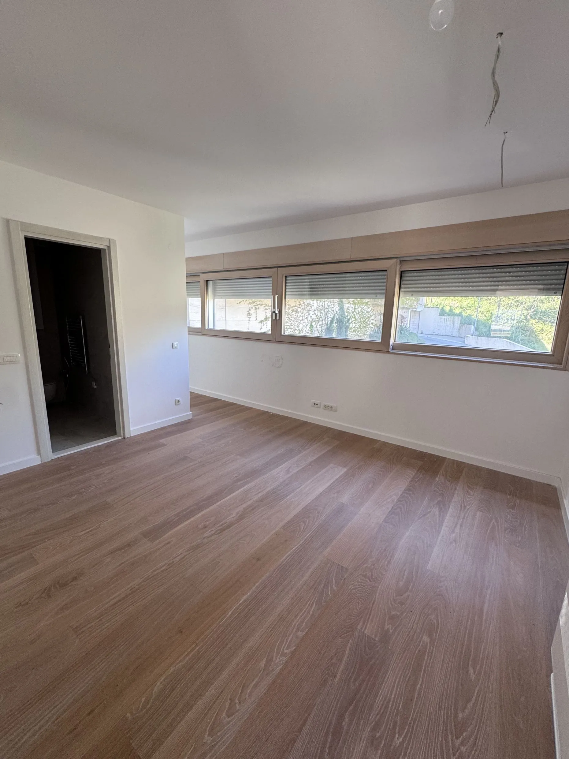 Leeres Zimmer mit Holzboden und großen Fenstern. Eine Tür zu einem anderen Raum ist sichtbar sowie Kabel an der Decke.
