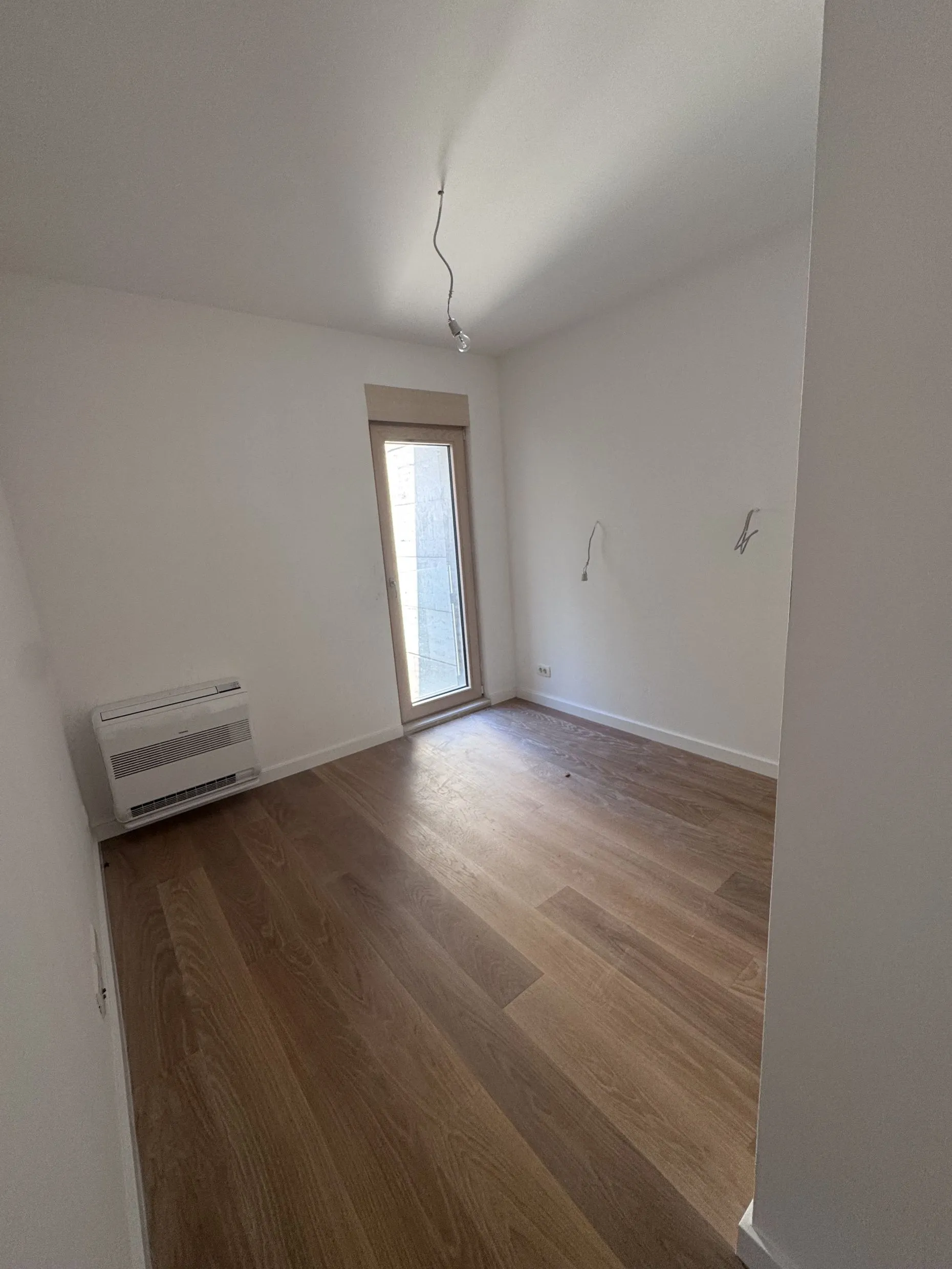 Leerer Raum mit Holzboden, weißen Wänden, Glastür und Wandklimaanlage.