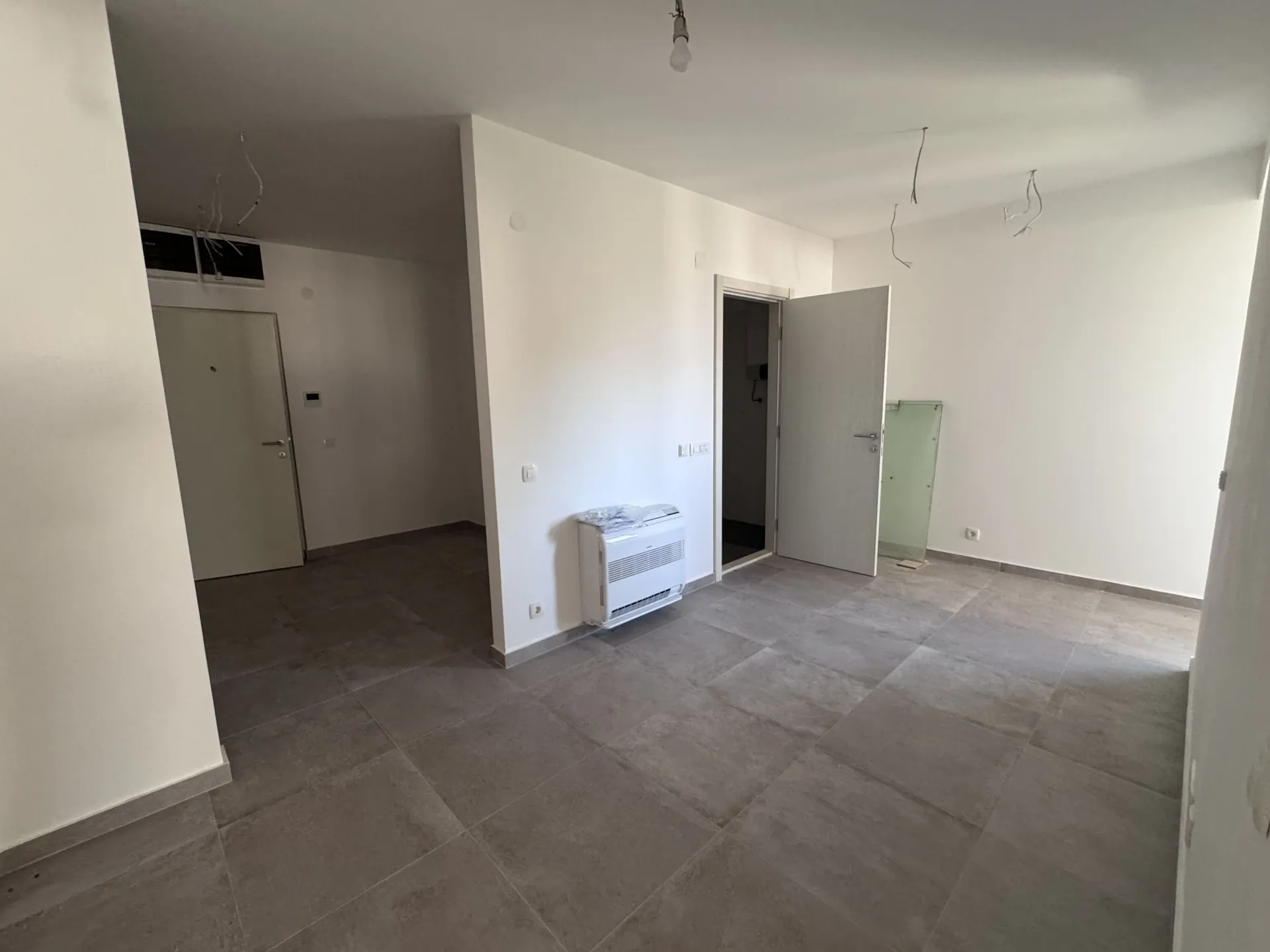 Leerer Raum mit grauen Fliesen und weißen Wänden. Offene Kabel an der Decke, ein weißer Konvektor und mehrere Türen sind sichtbar.