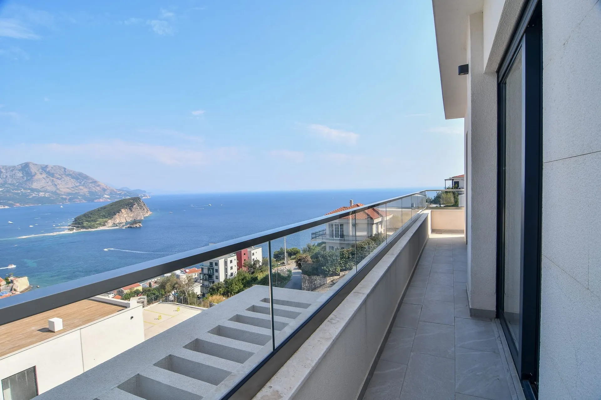 Spacieux balcon avec vue panoramique sur la mer et l’île à Budva, Monténégro : finition moderne et garde-corps en verre.