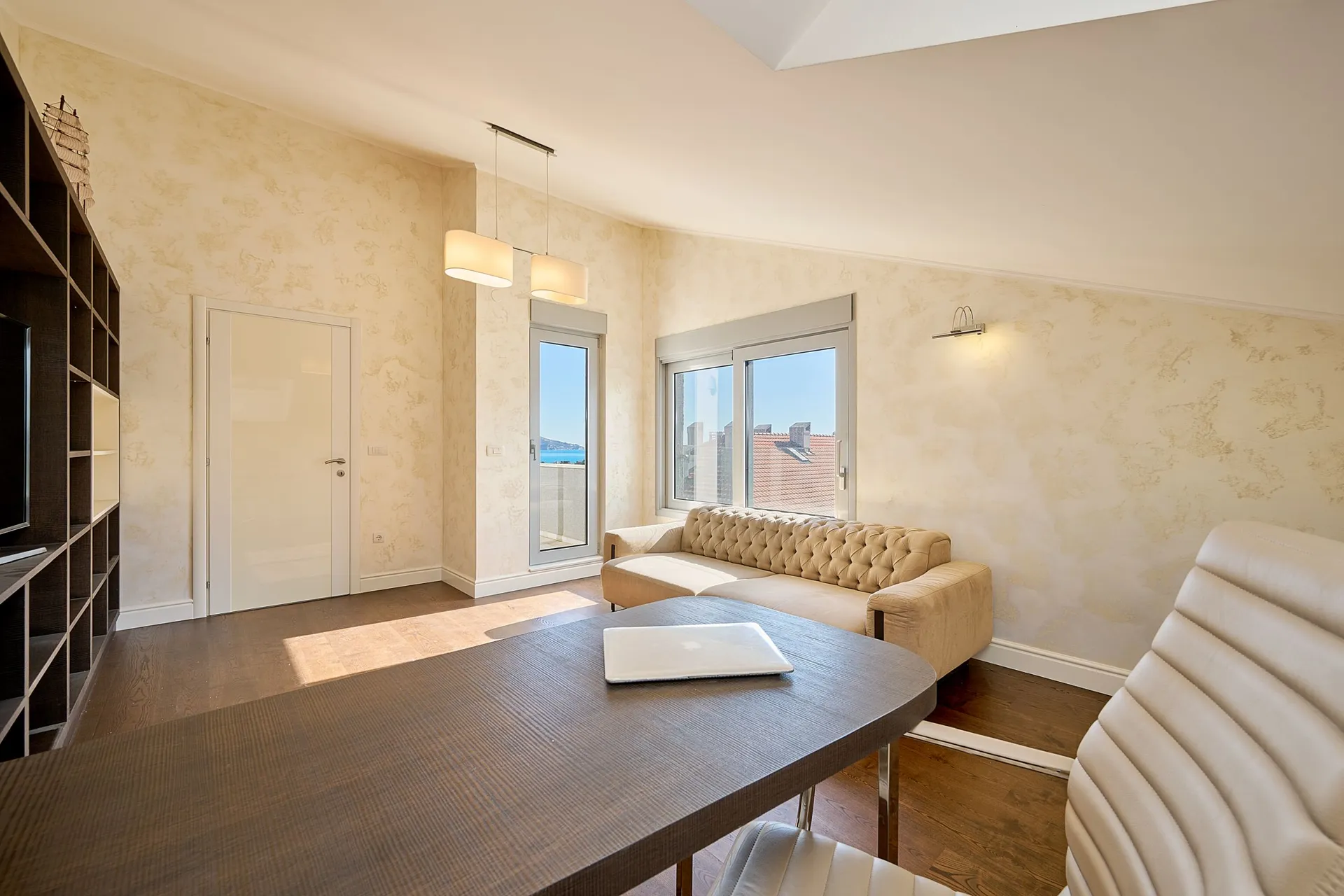 Heller Raum mit beigefarbenem Sofa, dunklem Tisch und Regal. Ein Laptop liegt auf dem Tisch vor Stadtblick.