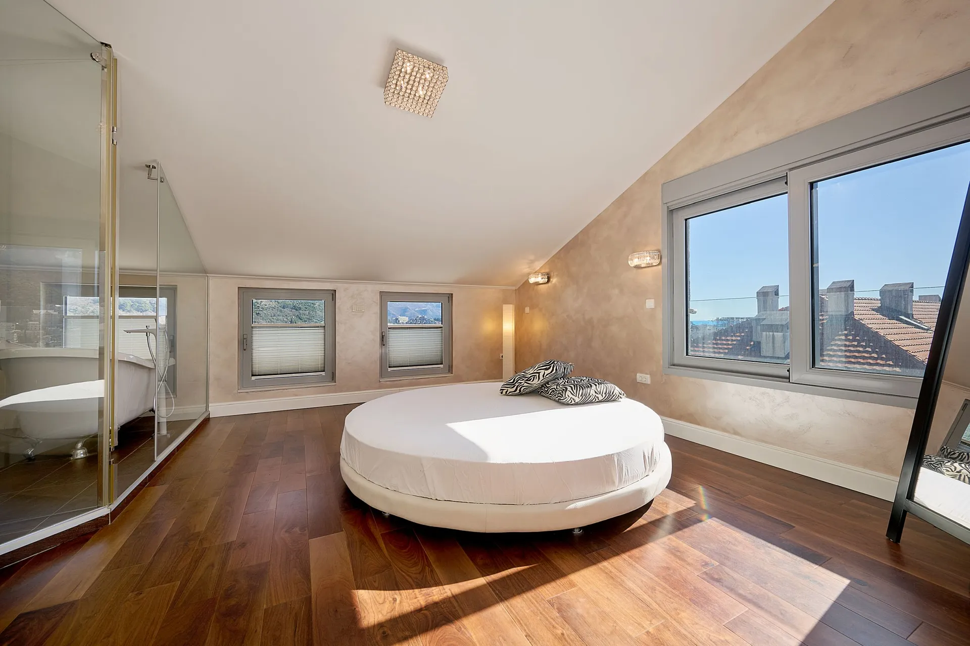 Moderne Schlafzimmer mit rundem Bett und Holzboden. Links zeigt eine Glastrennwand einen angrenzenden Badbereich.