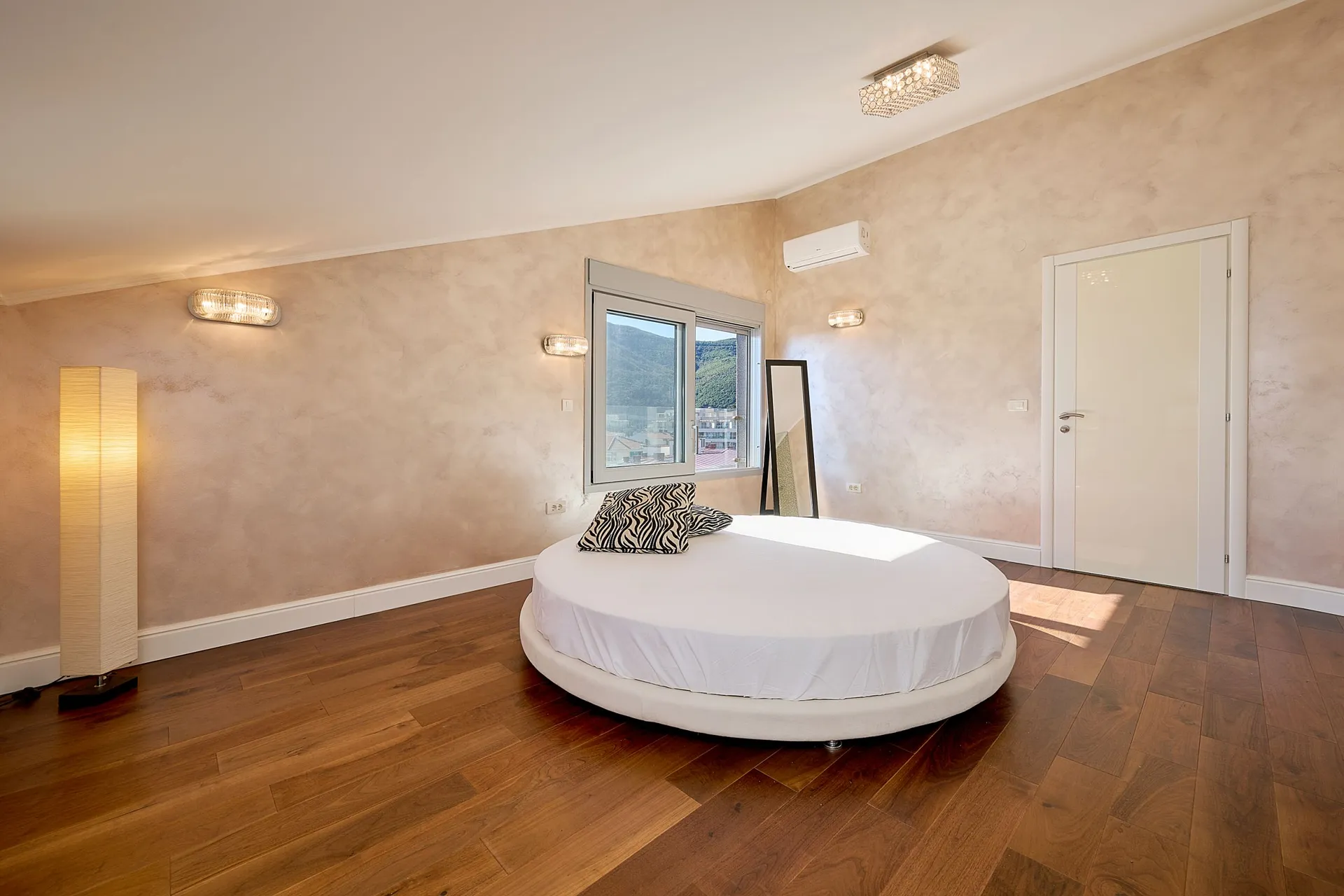 Moderne Schlafzimmer mit rundem weißen Bett, dunklem Holzboden und texturierten beigefarbenen Wänden.