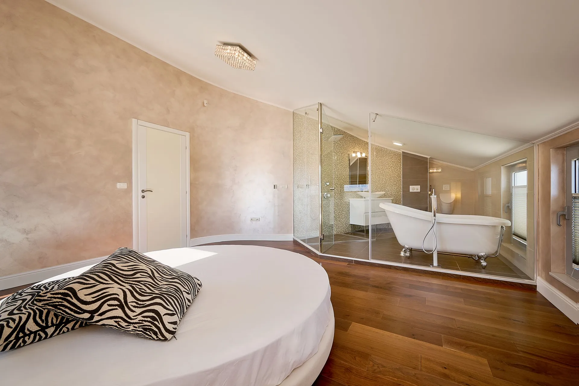 Schlafzimmer mit einem runden Bett und dekorativem Kissen. Eine angrenzende Badezimmer ist durch eine Glaswand mit einer Freistehbadewanne sichtbar.