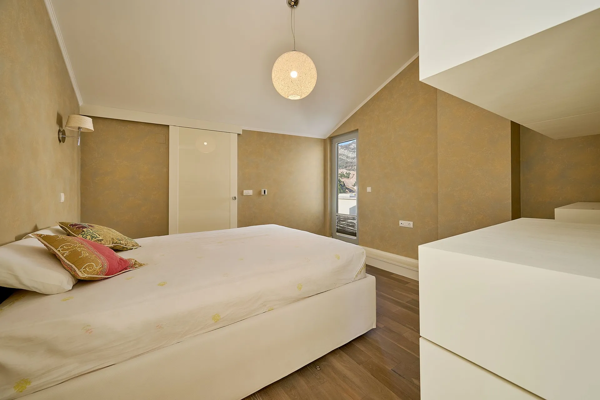 Moderne Schlafzimmer mit weißem Bett, goldfarbenen Wänden, einer runden Pendelleuchte und Holzboden.