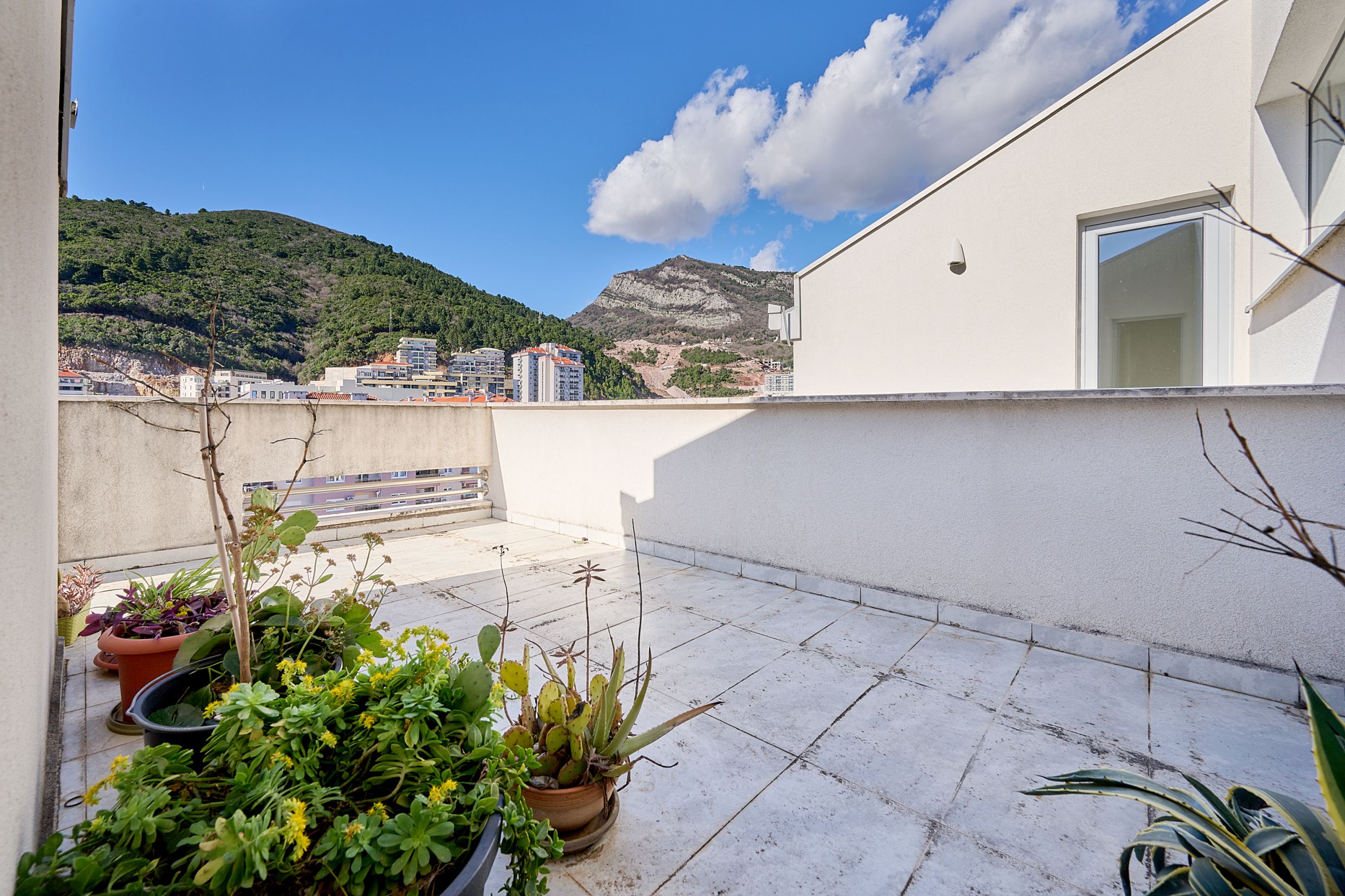 Prostrana terasa sa biljkama i pogledom na planine u stanu u Budvi, Crna Gora. Svetle pločice i otvoreni balkon.