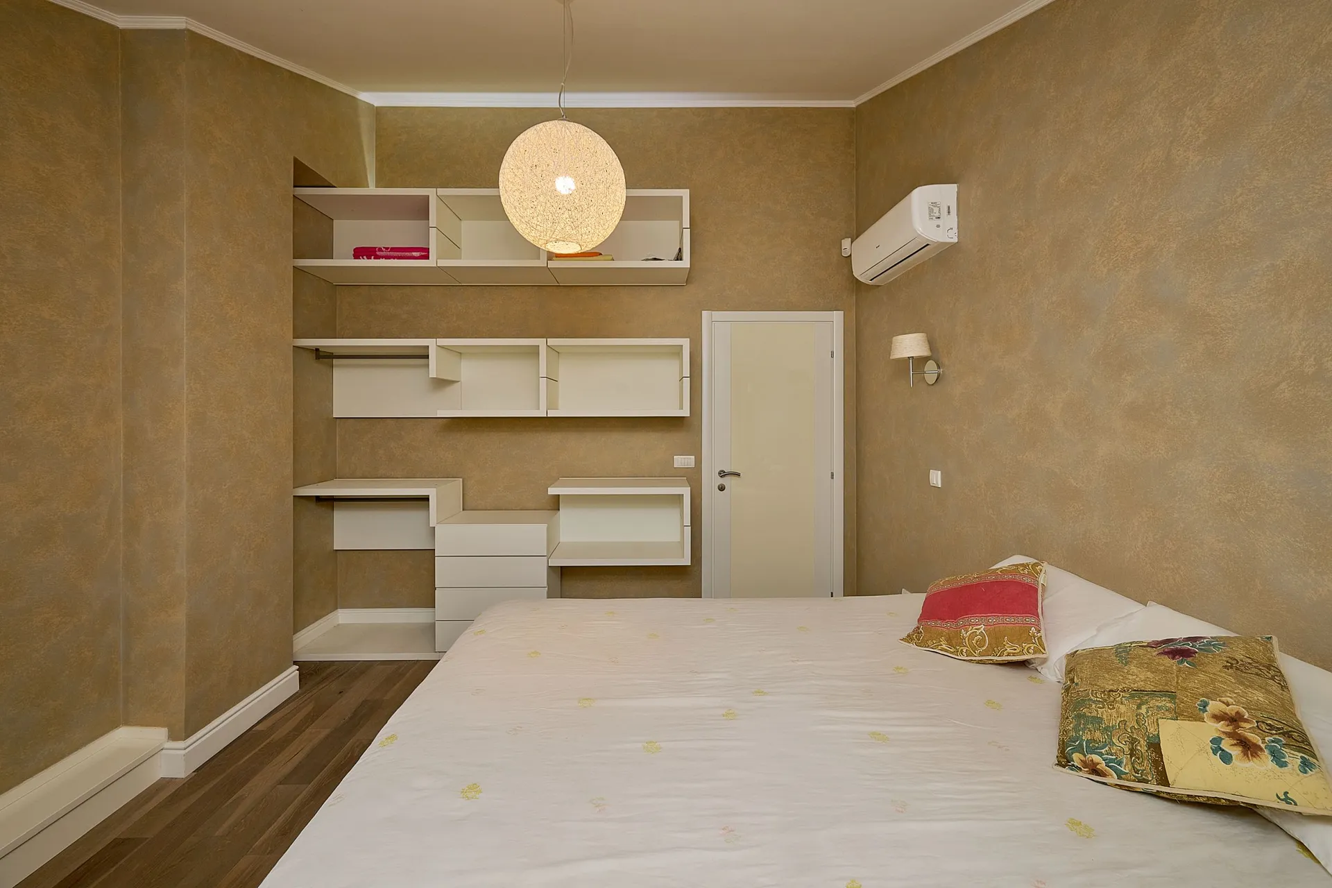 Moderne Schlafzimmer mit weißem Bett, eingebauten Regalen und einer runden Pendelleuchte vor beigefarbenen Wänden.