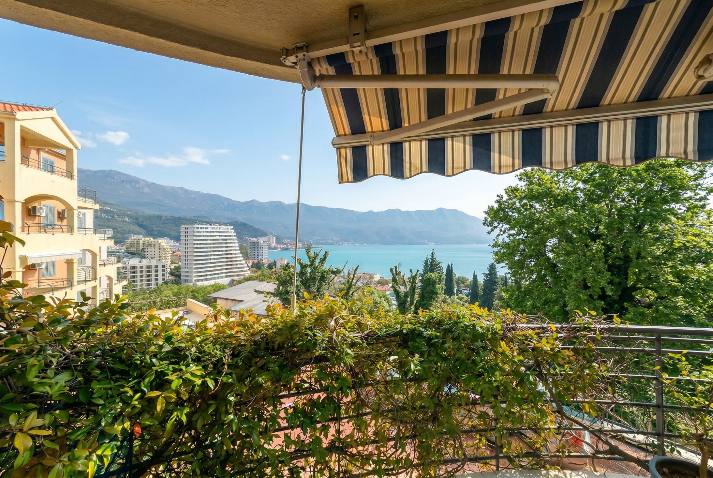 Blick vom Balkon einer Wohnung in Bečići, Montenegro: Panorama von Meer, Bergen und gestreifter Sonnenschutz über der Terrasse.