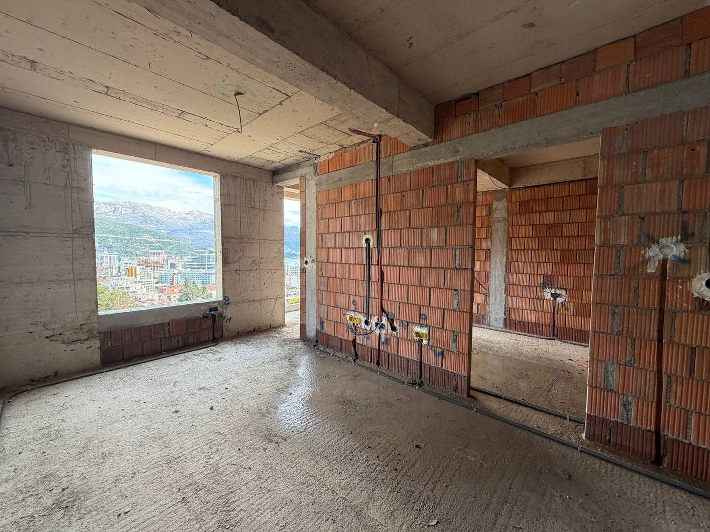Dnevna soba bez završne obrade u novogradnji u Budvi, Crna Gora: zidovi od cigle, betonski pod i panoramski pogled na grad.