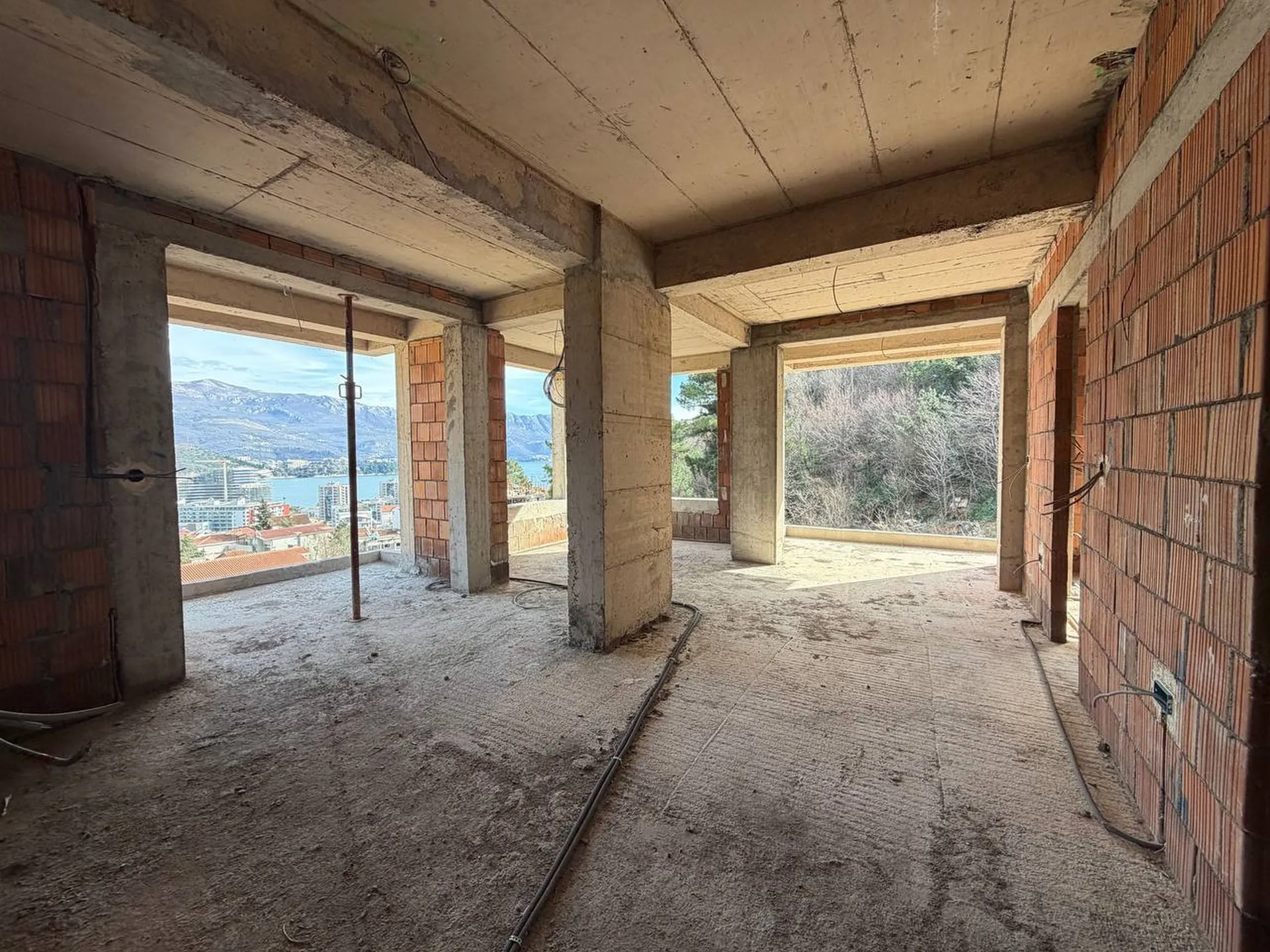Construction inachevée : piliers en béton, murs de brique et sol brut avec vue sur la ville et la mer.