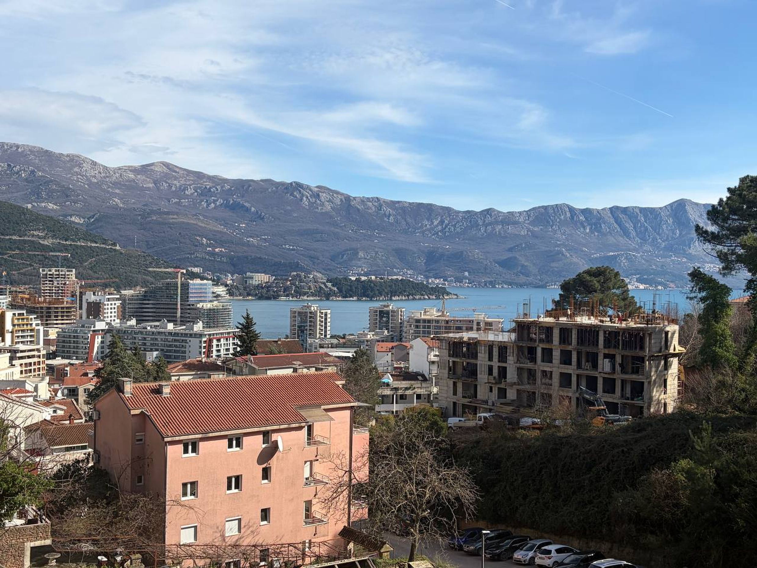 Panoramski pogled na grad Budvu, Crna Gora: stambene zgrade, objekti u izgradnji i morski zaliv u pozadini planinskih vrhova.