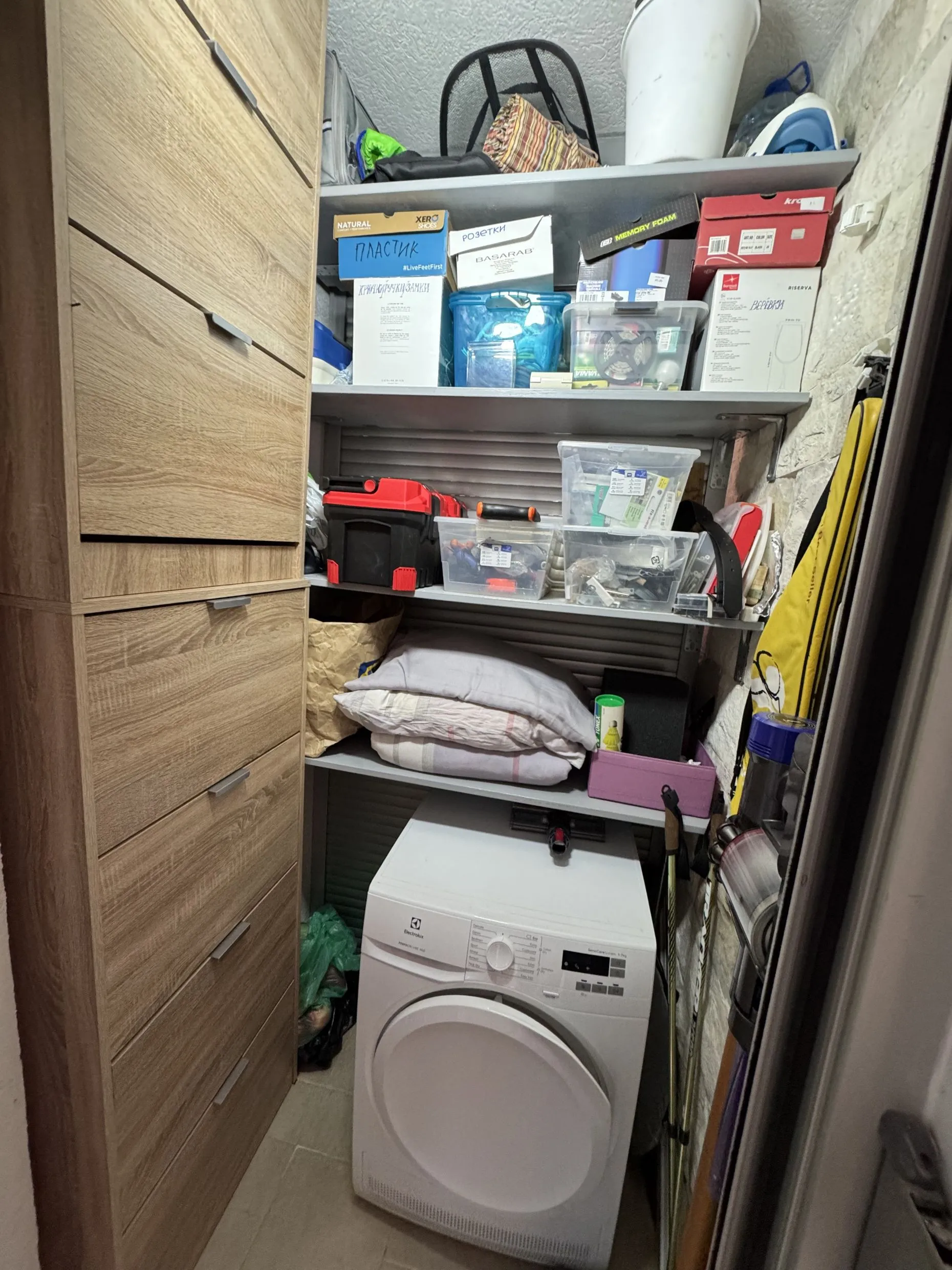 Small utility room featuring a wooden cabinet, washing machine, and shelves filled with boxes, tools, and various household items.