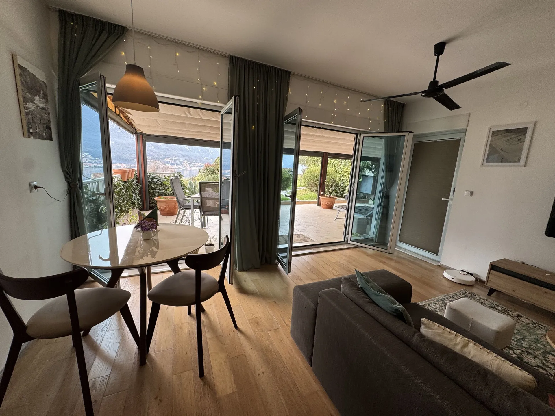 Bright living room with a grey sofa, round table, and open doors leading to a balcony overlooking a lake and mountains.