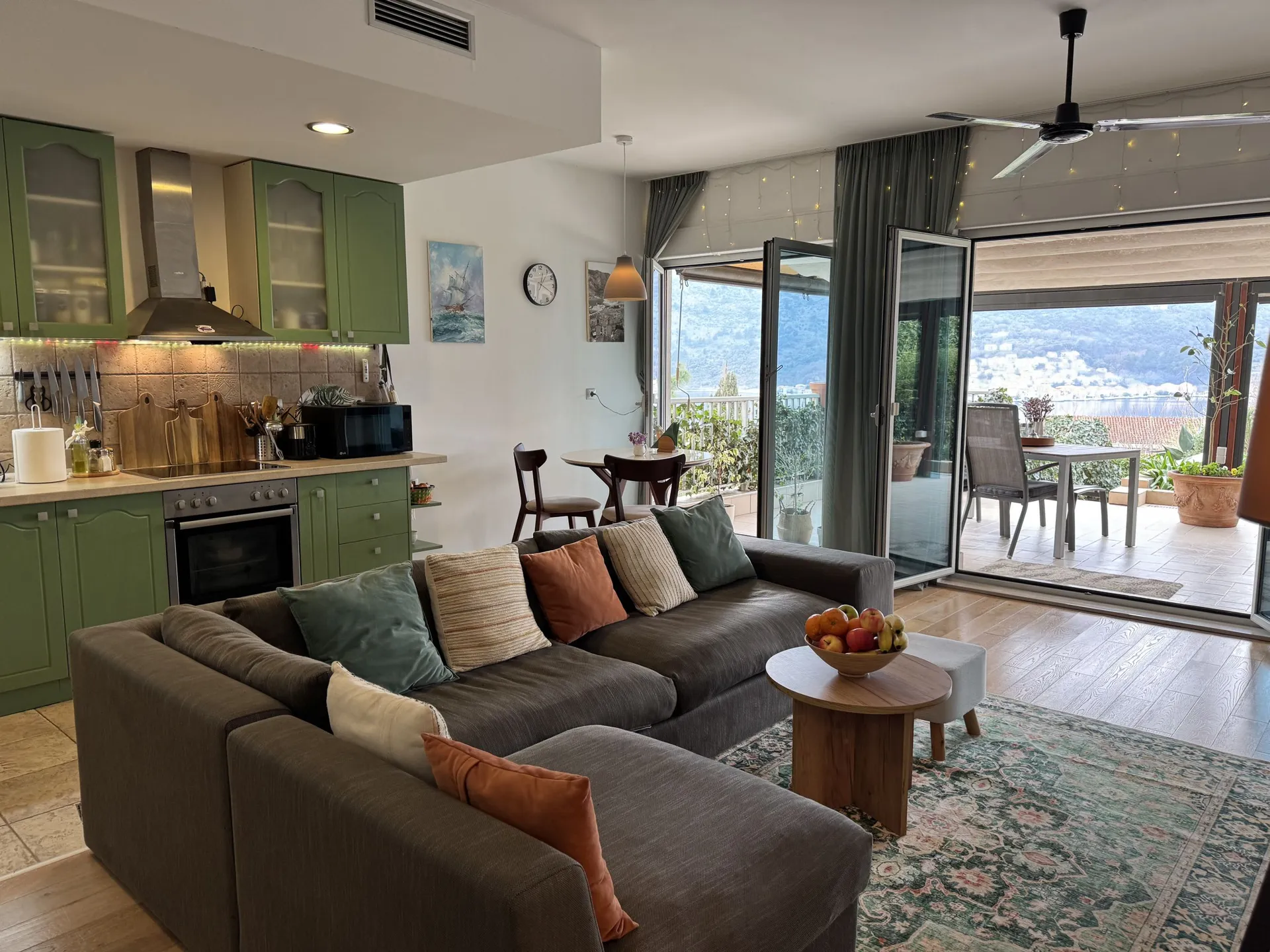 Living room with kitchen area, green cabinets and grey sofa. Sliding doors lead to a balcony with a view of water and mountains.