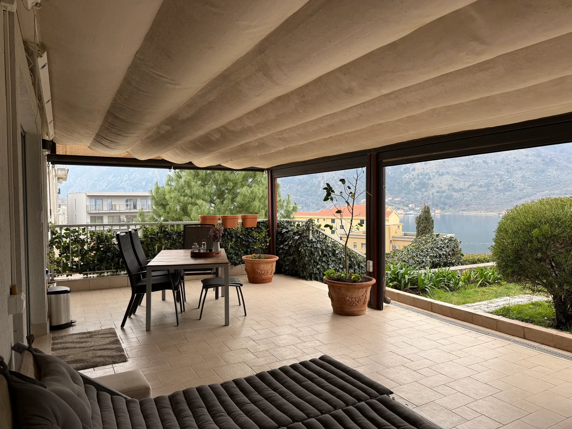 Terrace with dining table and sunbed under a canopy. View of the sea, mountains, and greenery.