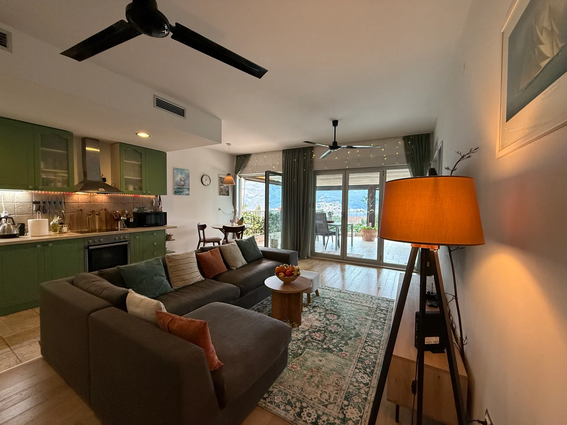 Living room with green kitchen, grey sofa, patterned rug and tripod lamp. Balcony with panoramic view visible through glass doors.