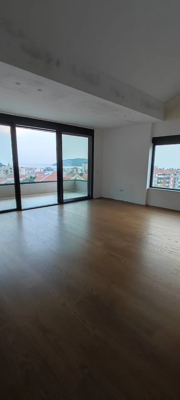 Ahşap zeminli ve büyük pencereli boş bir oda. Balkon kapısı ve dışarıda şehir manzarası görünüyor.