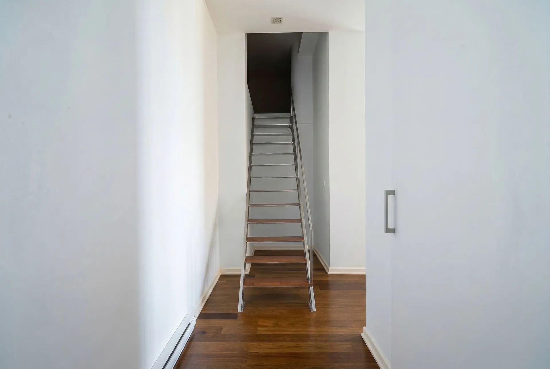Eine schmale weiße Treppe mit Holzstufen und Metallgriff führt über eine dunkle Öffnung nach oben.