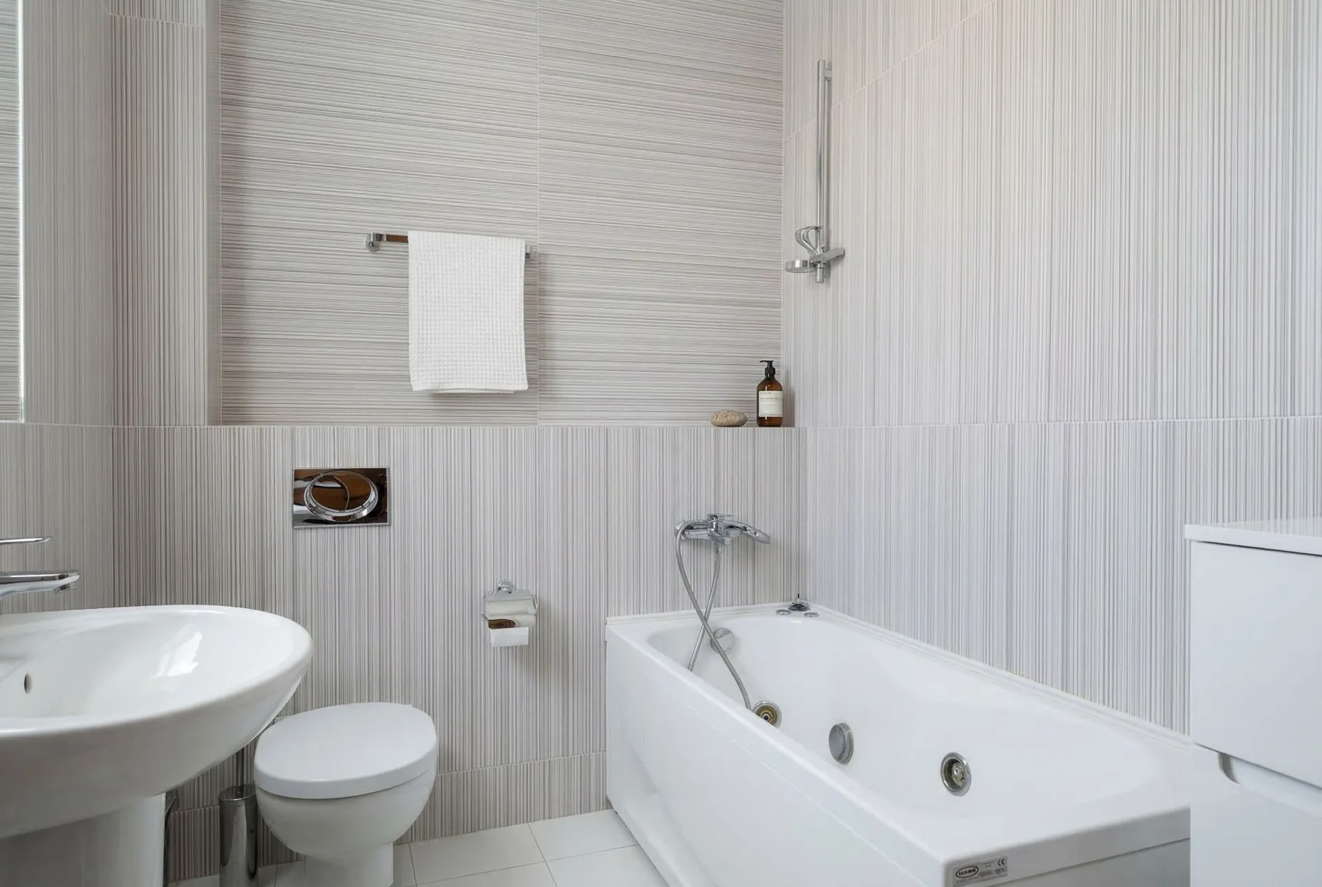 Moderne Badezimmer mit weißer Badewanne, Toilette und Spüle vor hellen strukturierten Fliesen.