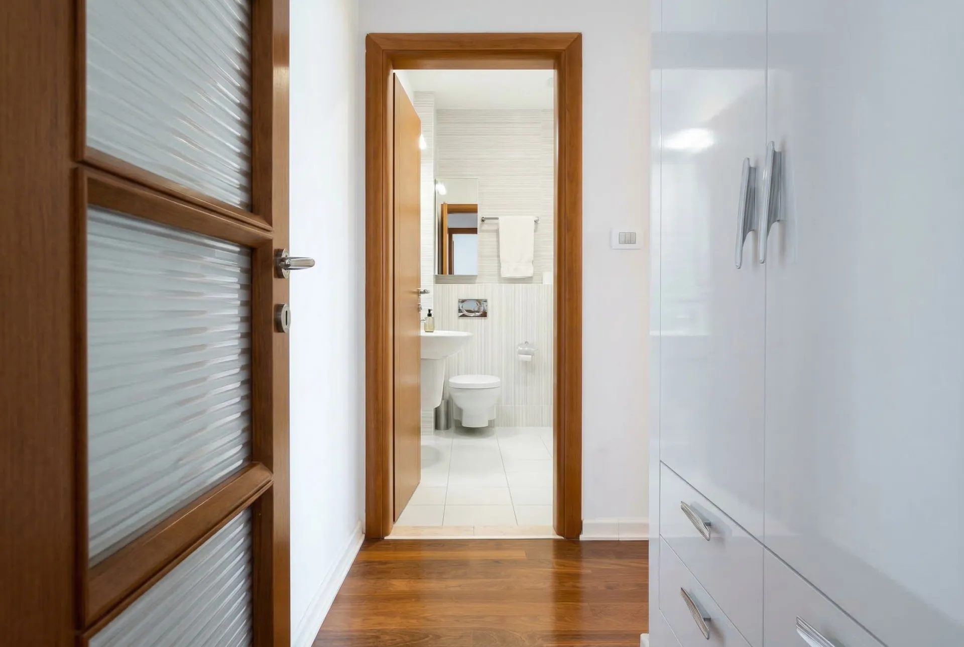 Blick aus einem Flur in ein weißes Badezimmer mit Toilette, Spüle und Handtuch an der Wand.