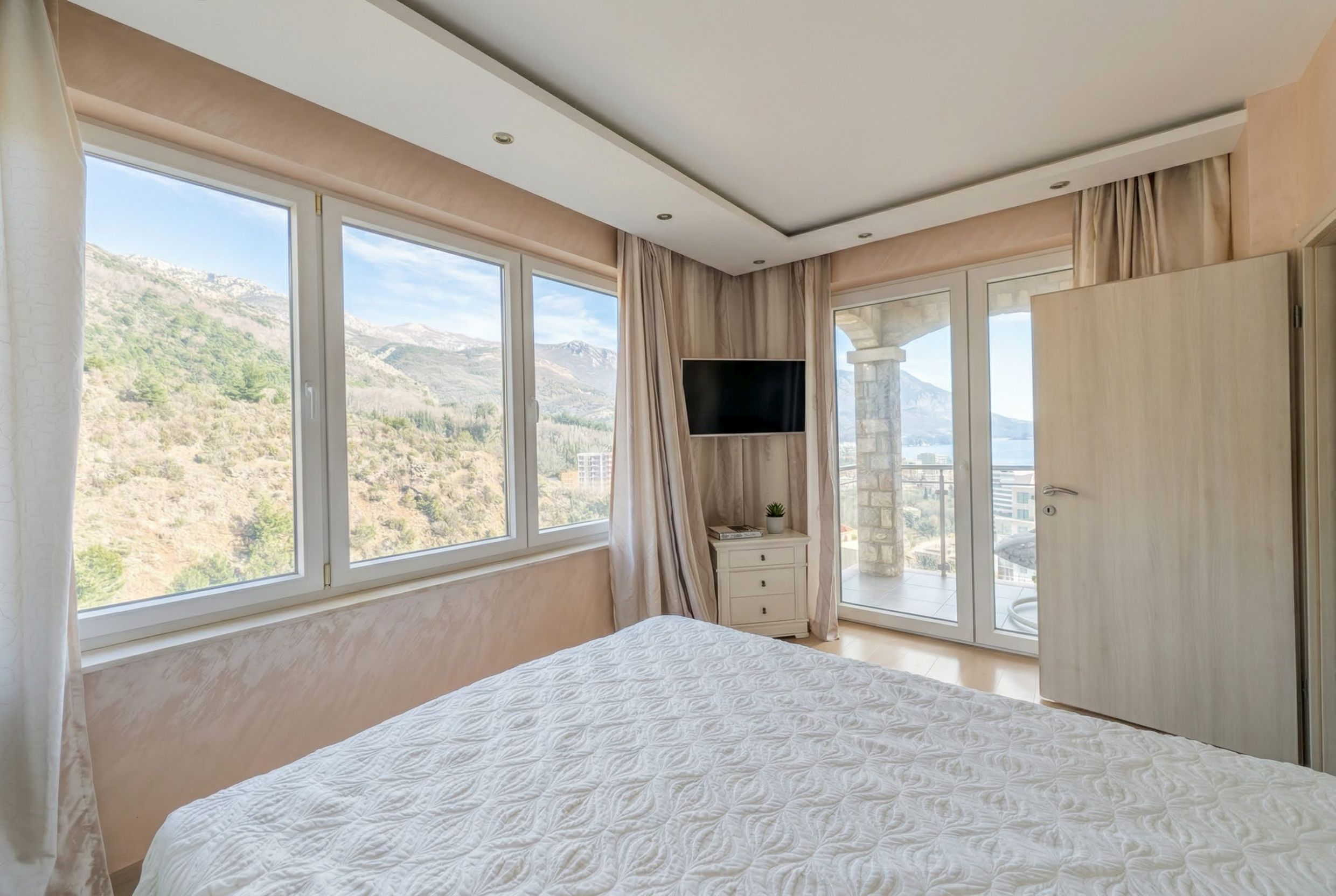 Bedroom in an apartment in Bečići, Montenegro: panoramic windows with mountain views, light-colored furniture, and access to the balcony.