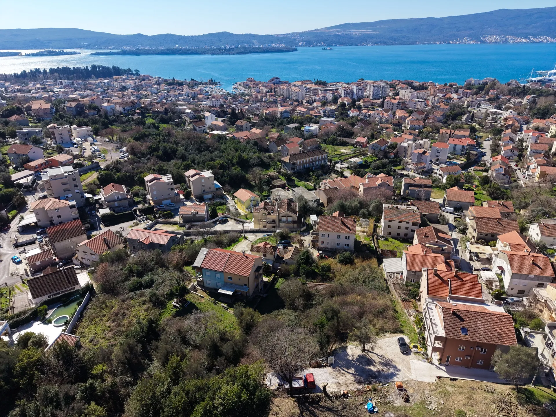 Kıyı şeridindeki, kırmızı kiremitli evlere sahip, yeşilliklerle çevrili ve mavi deniz ile dağlar arka planda olan bir şehrin havadan görünümü.