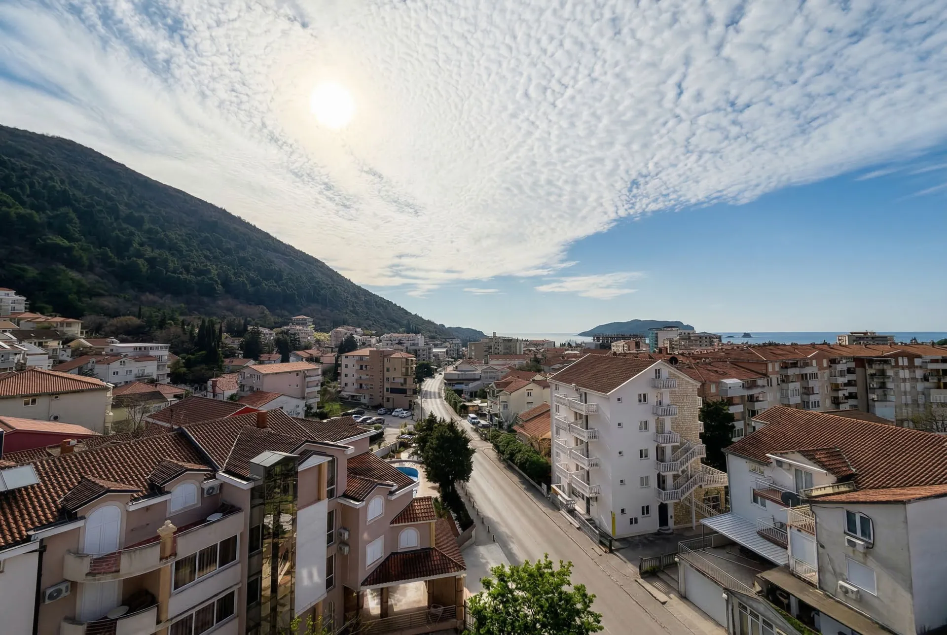 Kırmızı çatılı şehir evleri, bir yol, yeşil bir dağ yamacı ve deniz üzerine panoramik görünüm.