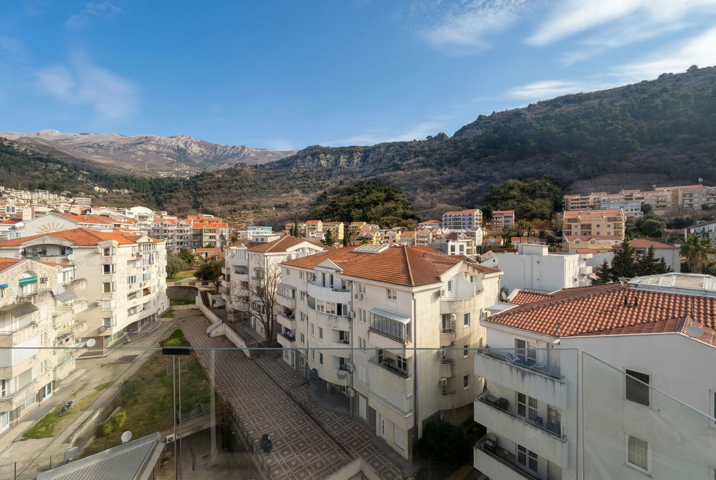 Blick vom Balkon eines Penthouses in Budva, Montenegro: Stadtpanorama, Wohnhäuser mit Ziegeldächern und Berglandschaft.