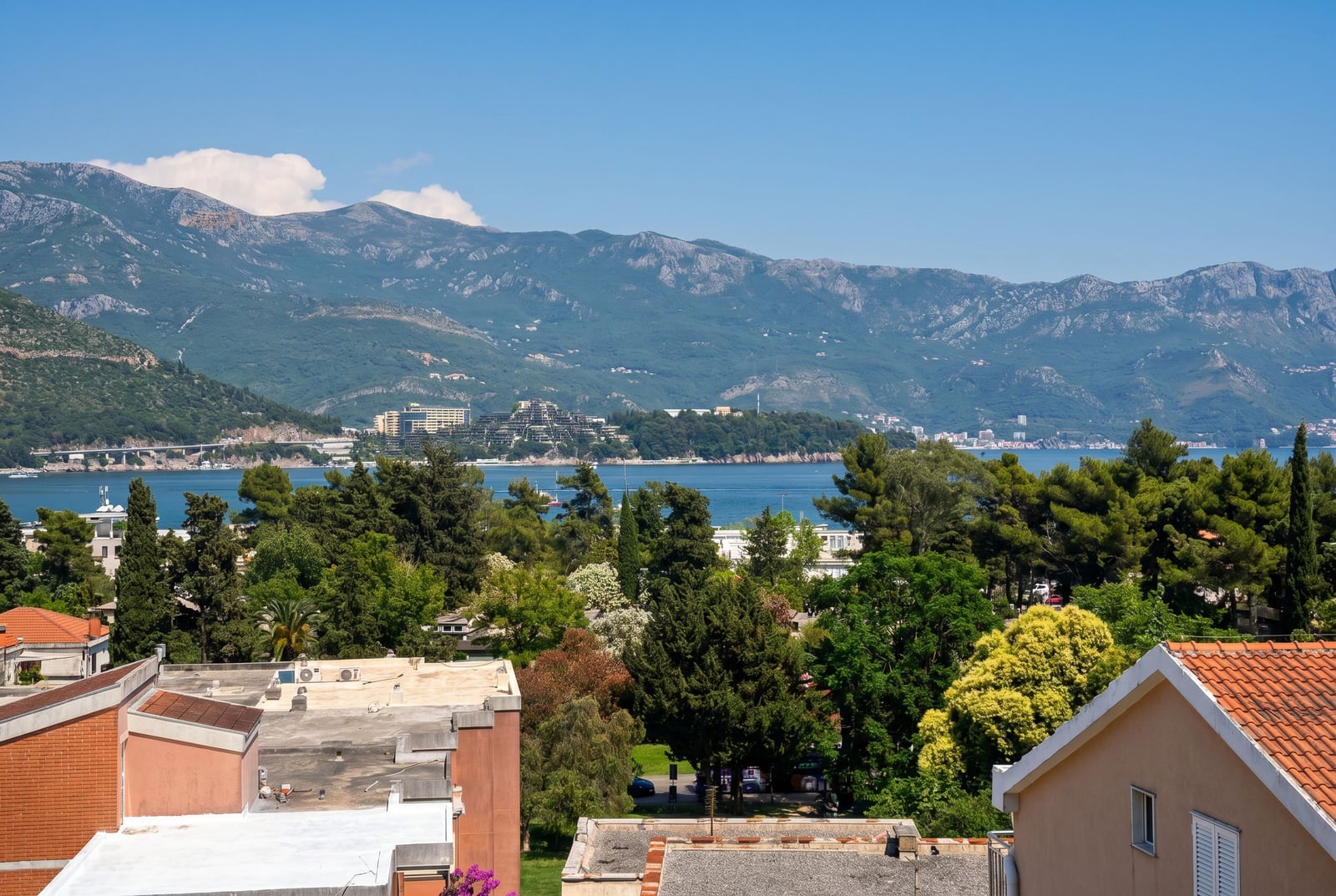 Panoramic view of the sea, mountains, and the resort town of Budva, Montenegro, from the window of an apartment with a terrace and a green area.