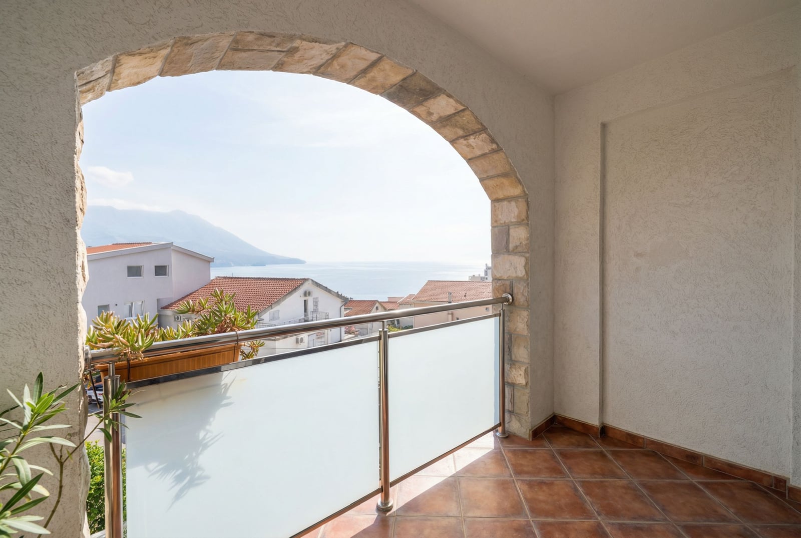 Balkon sa kamenim lukom i pogledom na more u hotelu u Bečićima, Crna Gora: pločice na podu i staklena ograda.