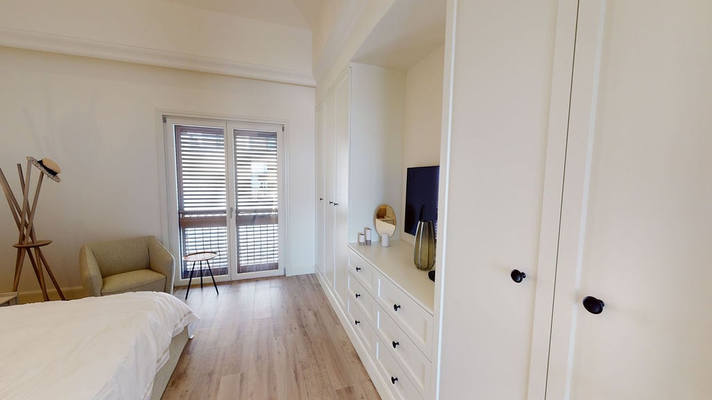 Svetla spavaća soba u stanu, Luštica, Crna Gora: ugradbeni ormar, komoda, fotelja i izlaz na balkon sa roletnama.