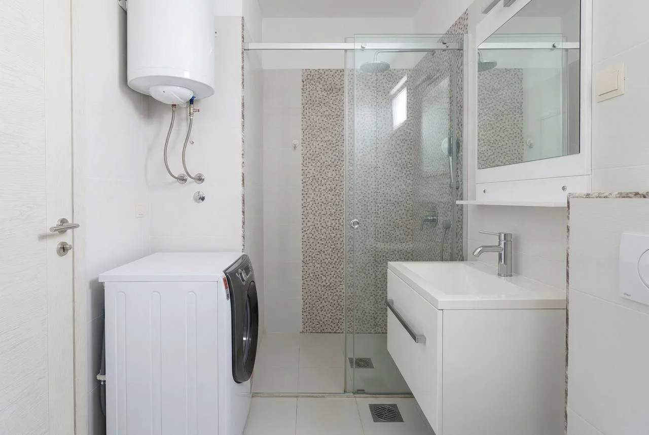 Cam duş, tezgah, çamaşır makinesi ve duvara monte su ısıtıcısı olan banyo.