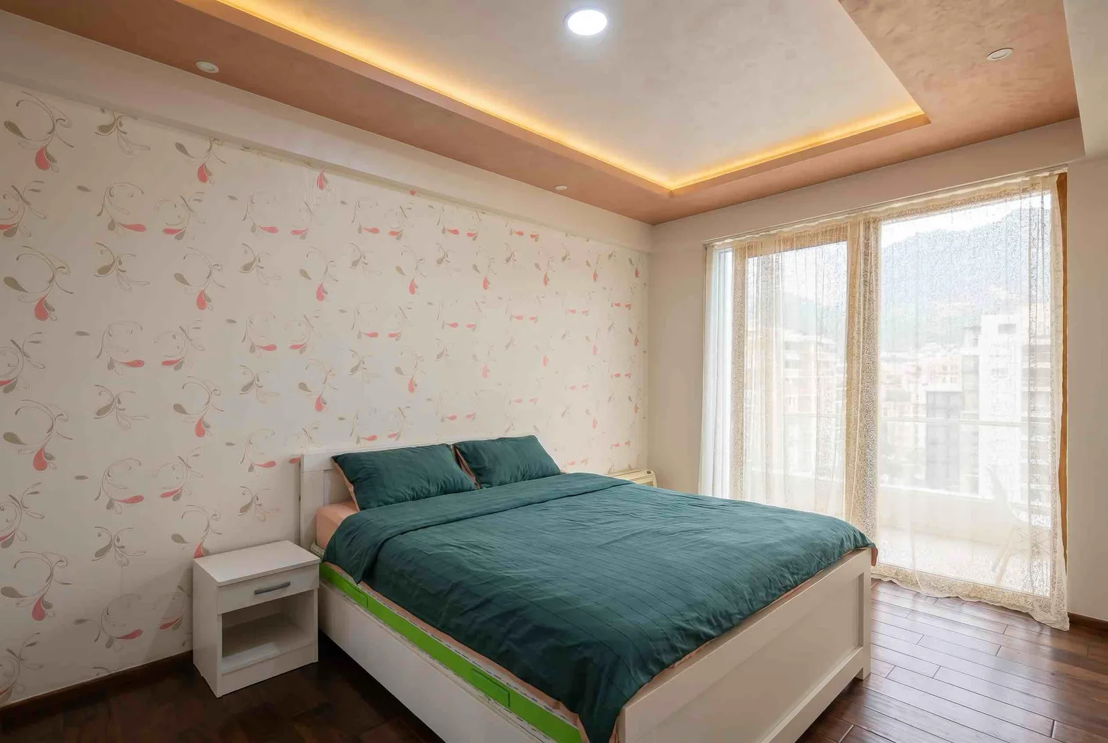 Bright bedroom featuring a double bed with green bedding, a white nightstand, floral wallpaper, and large windows with sheer curtains.