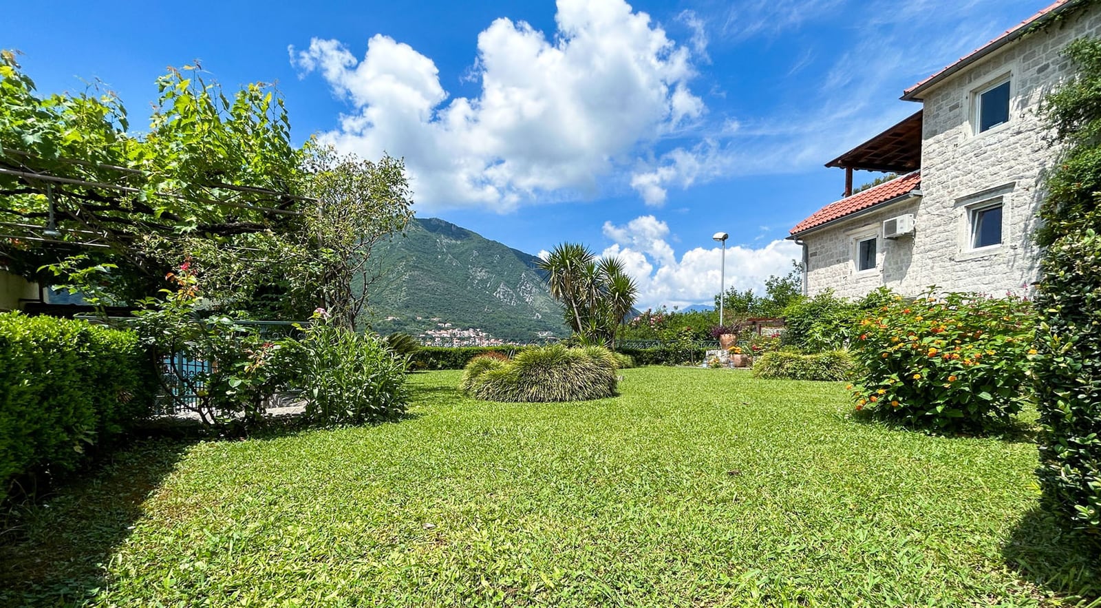Grüner Garten mit Steinvilla in Dobrota, Montenegro, mit Bergblick und Weinpergola auf dem Grundstück.