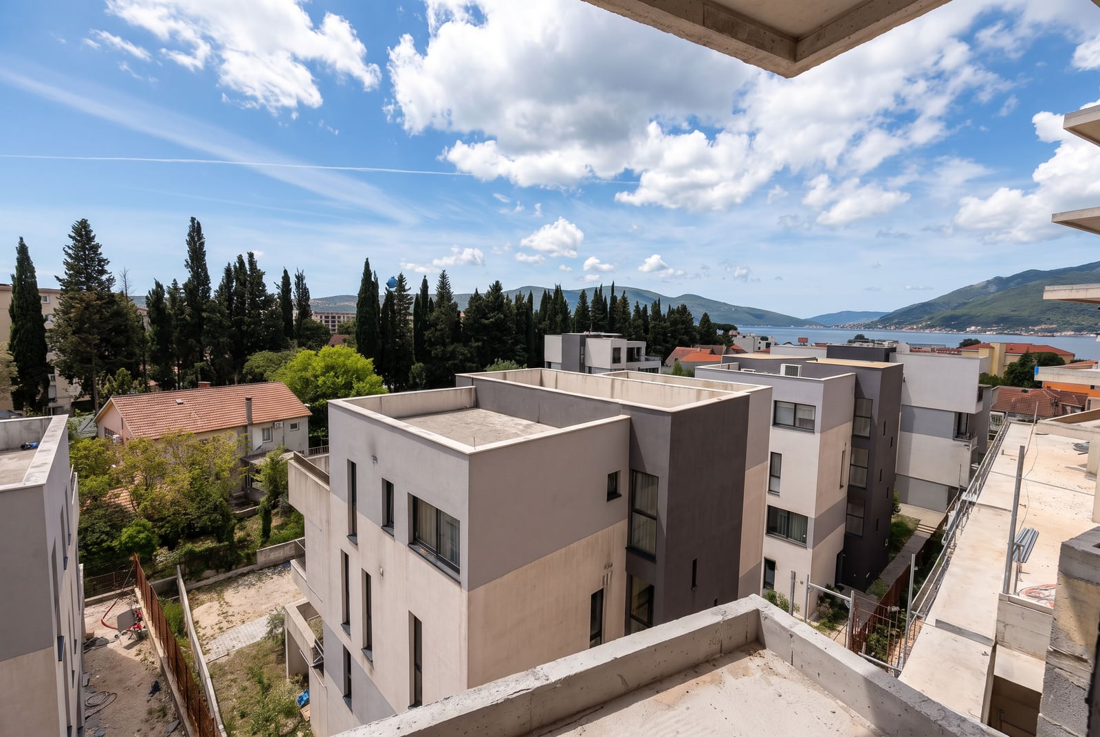 View from the balcony overlooking modern buildings, green trees, mountains, and the bay in Tivat, Montenegro.