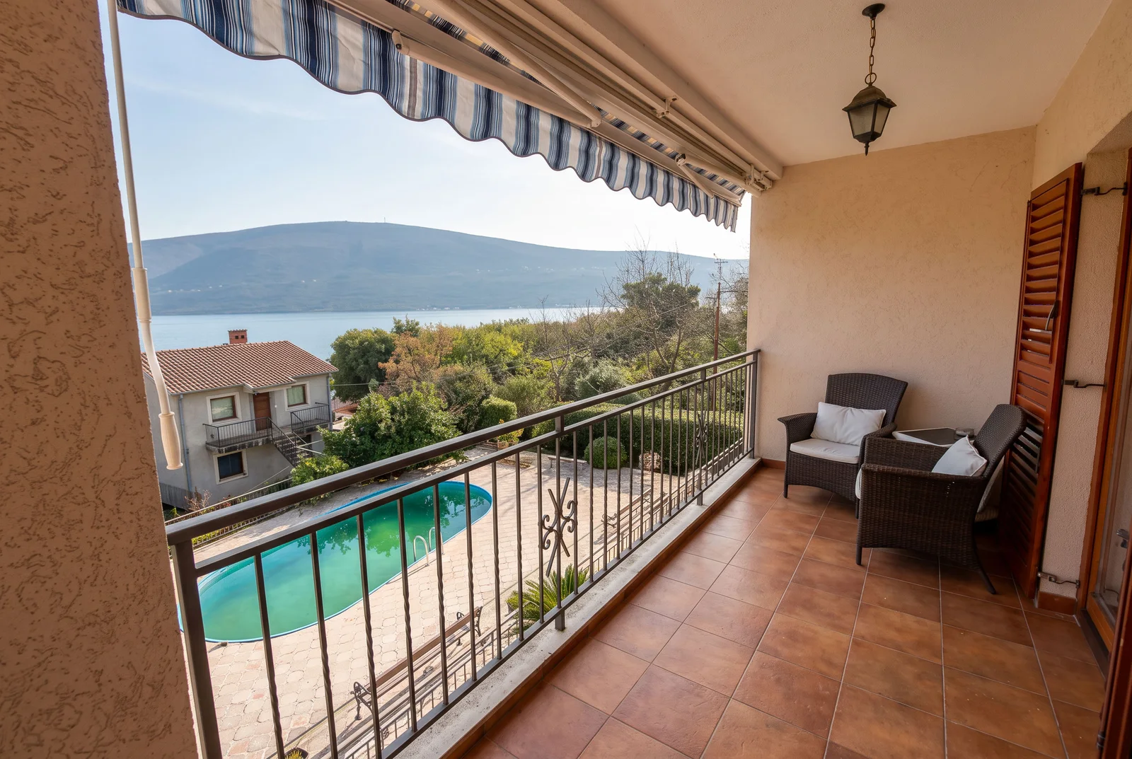 Balcon avec chaises en rotin et auvent rayé, vue sur piscine et lac.