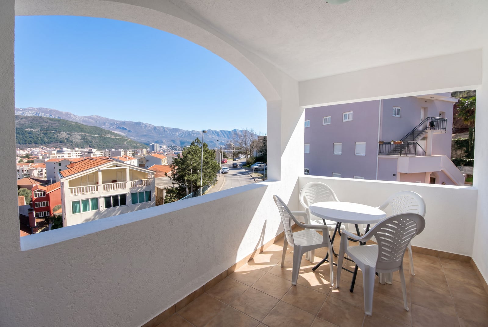 Prostran balkon sa belom plastičnom nameštajem i panoramskim pogledom na planine i grad u Budvi, Crna Gora