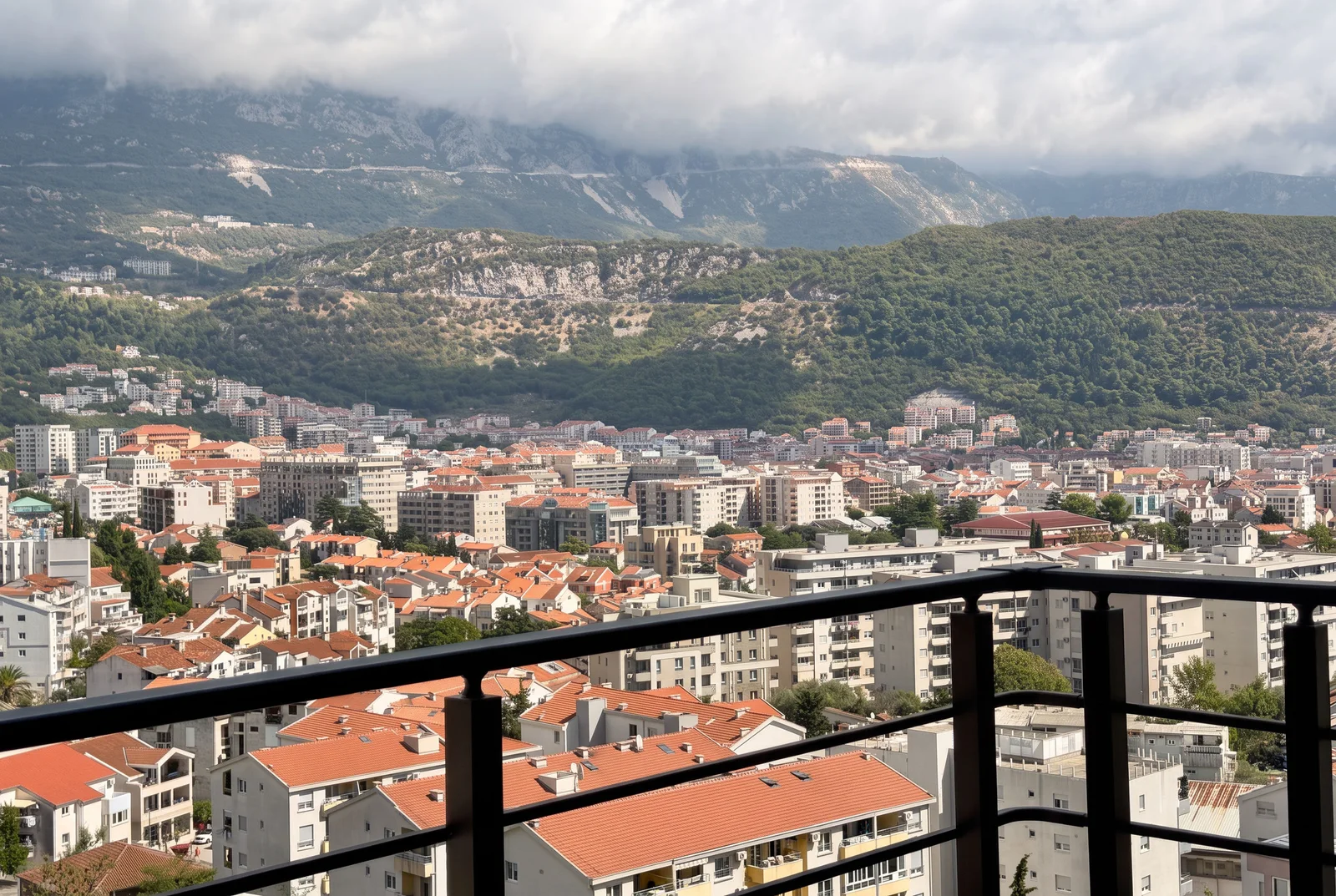 Pogled sa balkona na grad sa crvenim krovovima i planinskim masivom u pozadini.