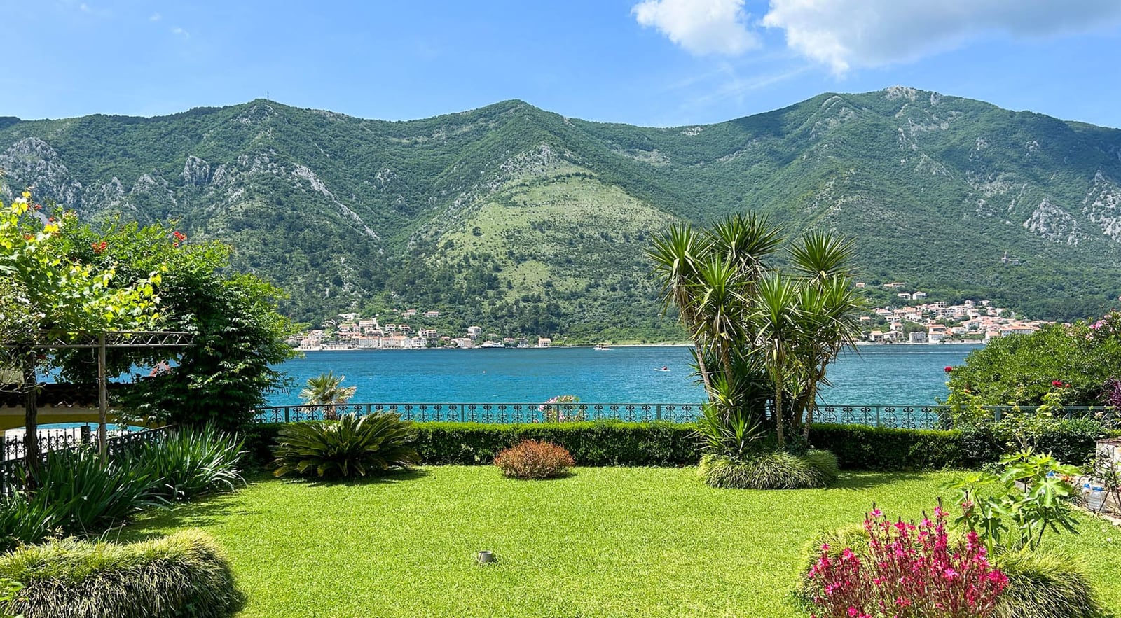 Blick auf das Meer und die Berge aus dem grünen Garten der Villa in Dobrota, Montenegro: gepflegter Rasen, Palmen und ein panoramischer Meerblick.