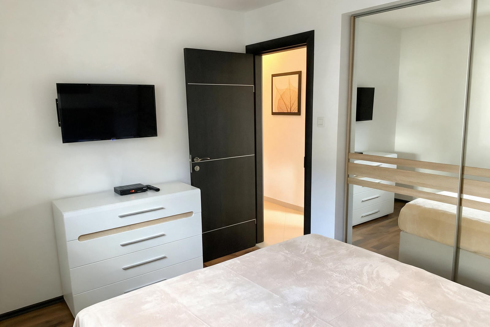 Bedroom with a white dresser, TV on the wall, and mirrored wardrobe in an apartment in Budva, Montenegro.