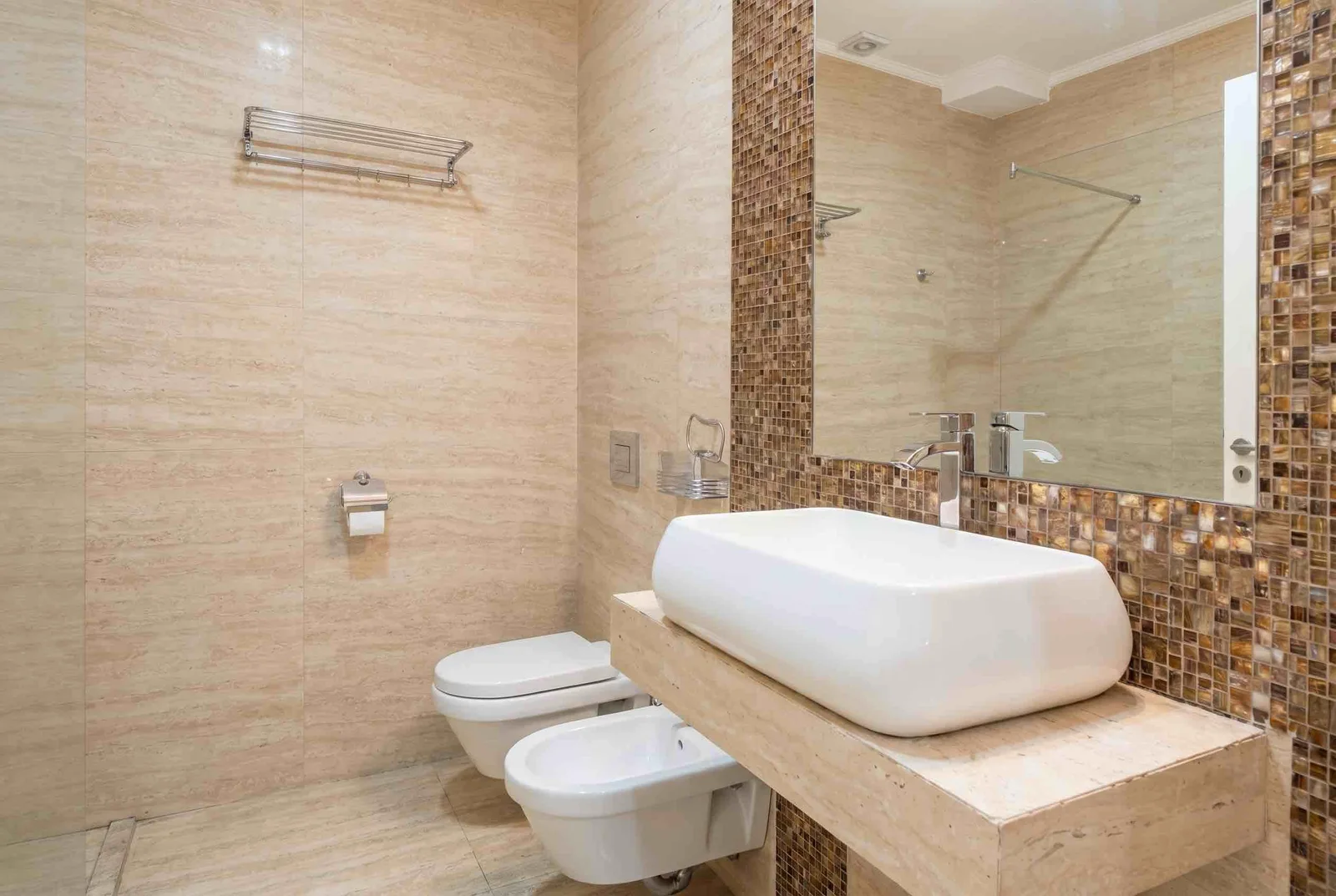 Modern bathroom with beige tiles, wall-mounted toilet, bidet, and a white rectangular vessel sink on a stone countertop next to a large mirror.
