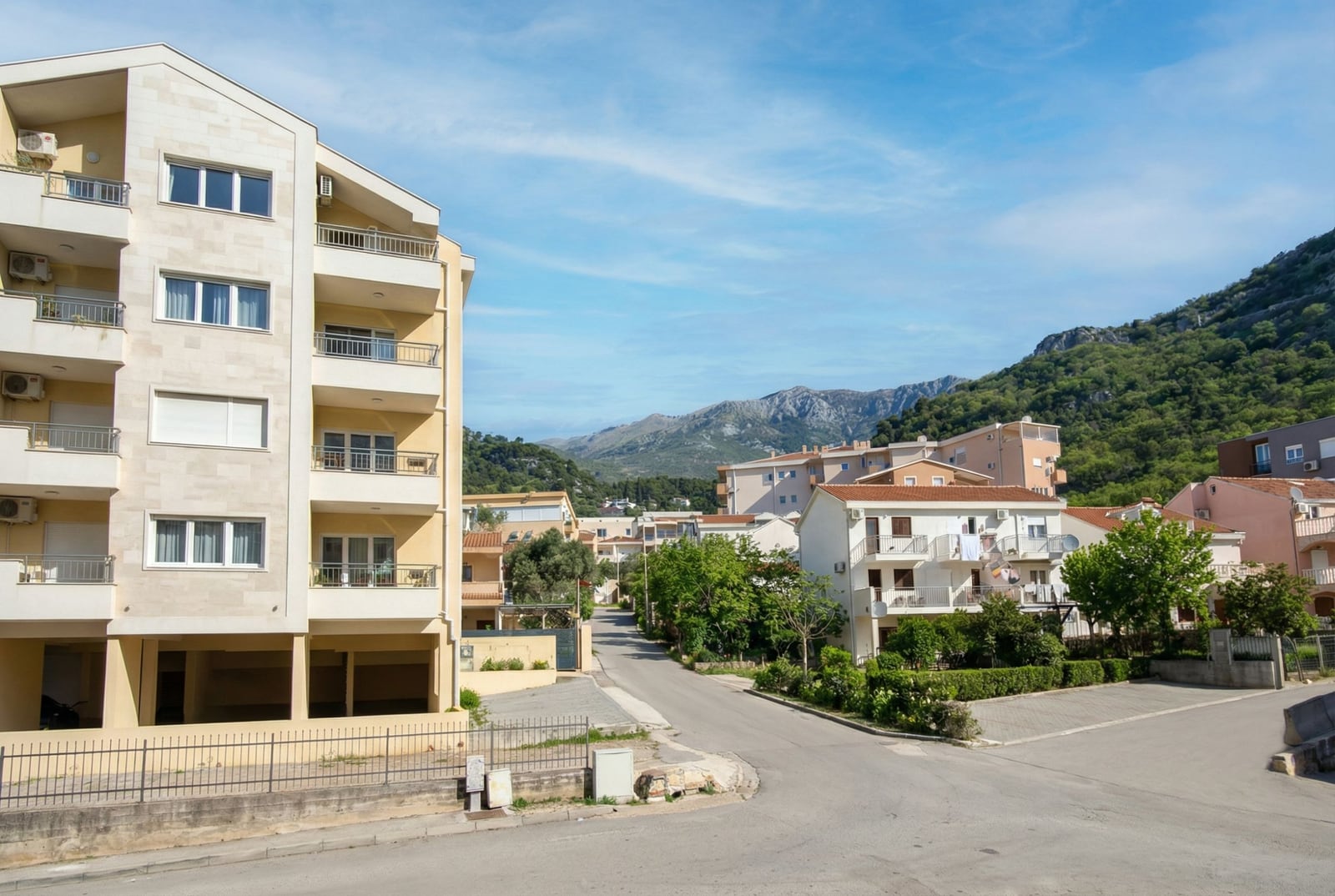 Savremeni stambeni kompleks u Budvi, Crna Gora: pogled na višespratnicu, ulicu i planinski pejzaž tokom sunčanog dana.