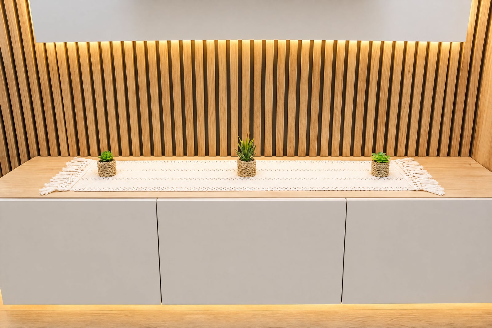 Modern wooden slat wall with warm lighting above grey cabinets featuring small plants on a white macrame runner.