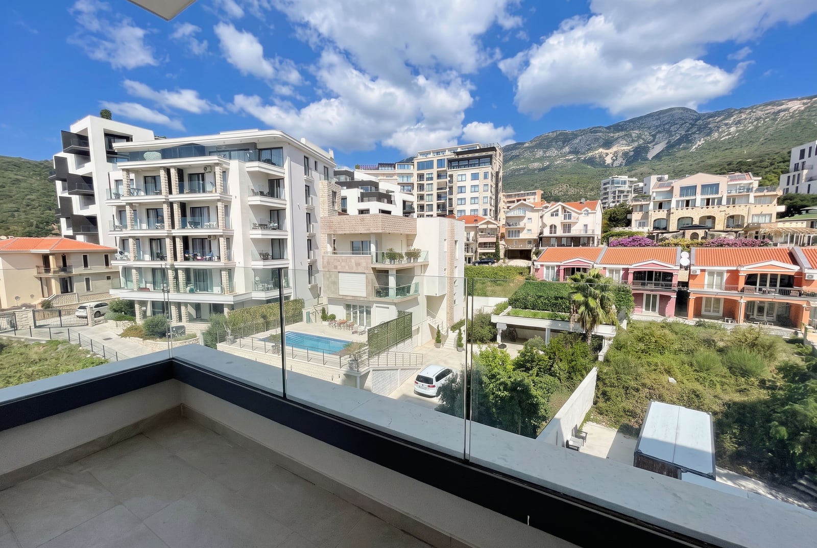 View of modern residential complexes, mountains, and a pool from the balcony of an apartment in Becici, Montenegro