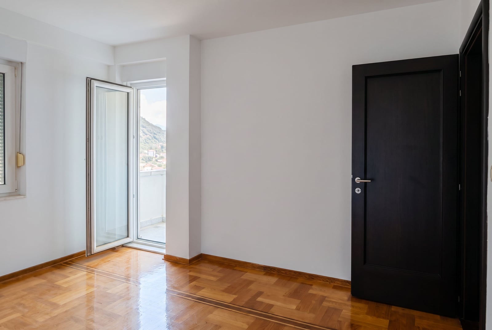 Bright empty bedroom with parquet flooring, a black door, and access to a balcony in an apartment in Petrovac, Montenegro.